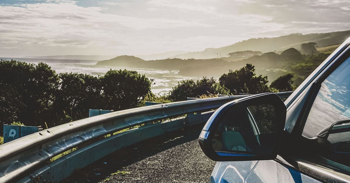 A car drives along a road with the ocean visible in the background under a clear blue sky.