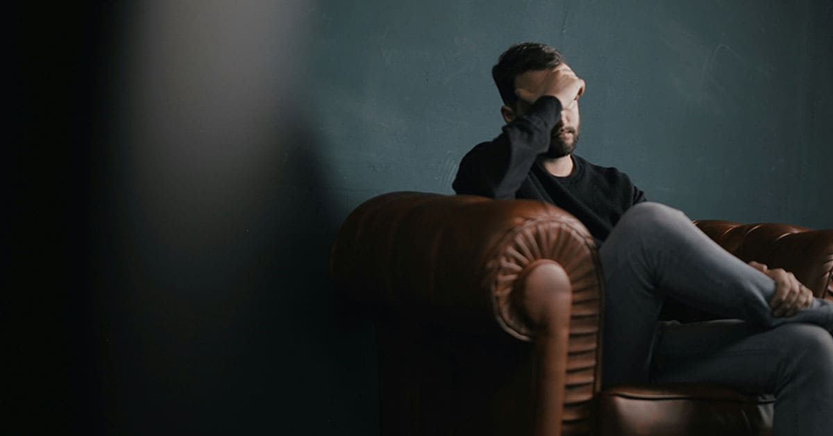 A man sitting on a leather chair against a black wall, looking relaxed and contemplative.