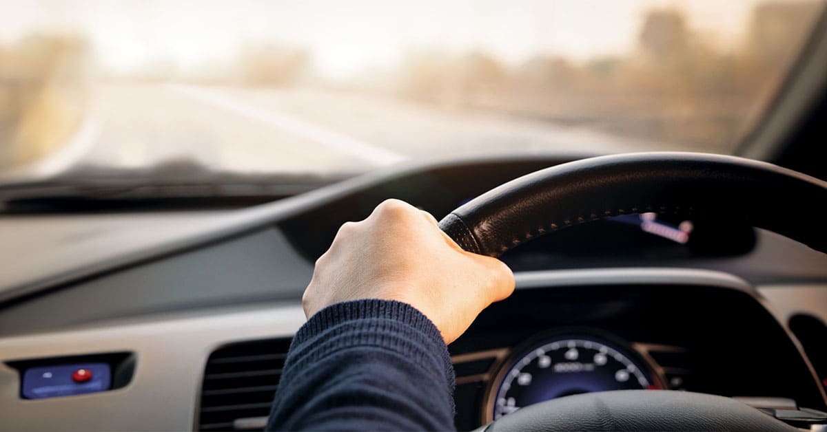 A person driving a car, gripping the steering wheel with one hand, focused on the road ahead.