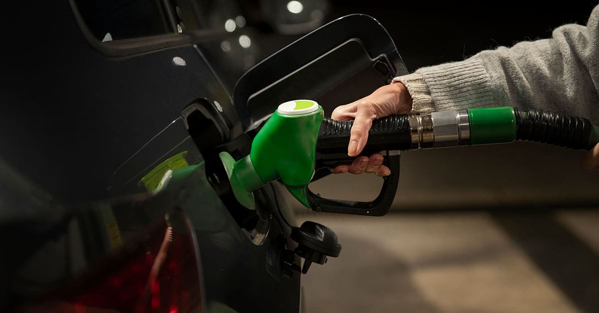 A person refuels their car at a gas station, holding the fuel nozzle and standing next to the car.