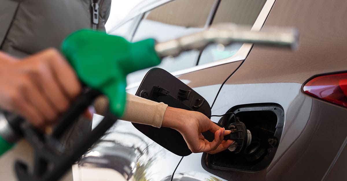 A person refuels their car at a gas station, holding the fuel nozzle and standing next to the vehicle.