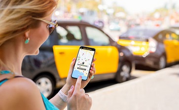 A woman checks a taxi app on her cell phone while standing on a city street. globalblog'