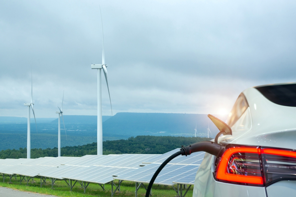 An EV charging overlooking solar panels and wind turbines in a lush green countryside