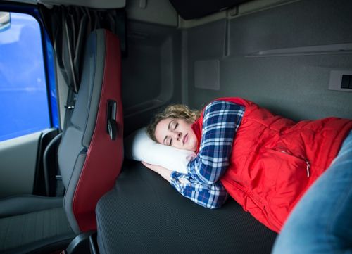A driver wearing warm clothes sleeps in the cab bed of their lorry
