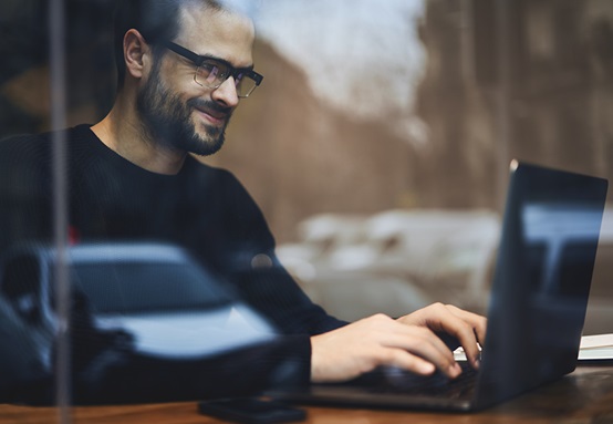 A person working on a laptop at a wooden table, viewed through a window that reflects parked vehicles and an urban street—symbolizing connectivity, mobility, and modern fleet management solutions in New Zealand.