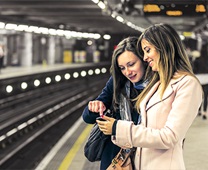 Two people on a subway platform, looking at an item in hand, with tracks and other commuters in the background.