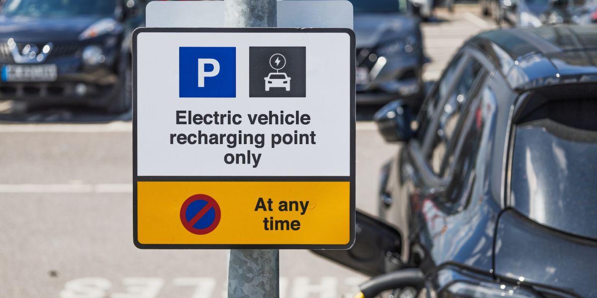 2026 budget salary sacrifice scheme impact: A UK car park sign reading “Electric vehicle recharging point only” and “At any time” with a no parking symbol, beside a dark grey electric car plugged in to charge.