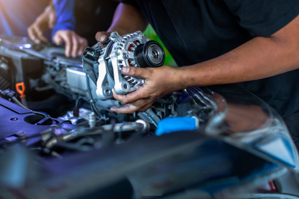 Cost of driving electric cars: A mechanic's hands holding an alternator over an open car engine bay, with another person working in the background.