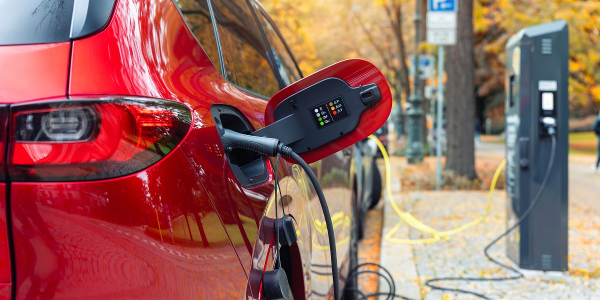 EV charging without a driveway ev charging without a driveway: A red car plugged into a public charging point by a black cable, parked on a tree-lined street with autumn leaves on the pavement.