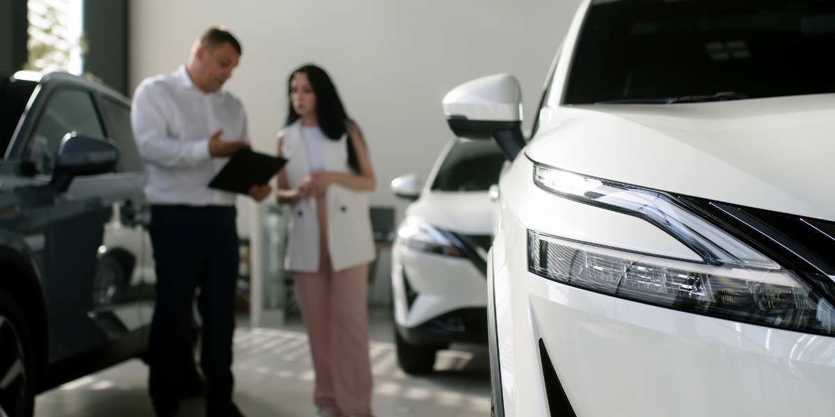 EV fleet transition plan ev fleet transition plan: A manager with a clipboard talking to a woman in a car showroom, with a white SUV's headlight in focus in the foreground.