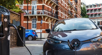 Electric car plugged into a residential street charging station in front of red-brick housing in an urban area.