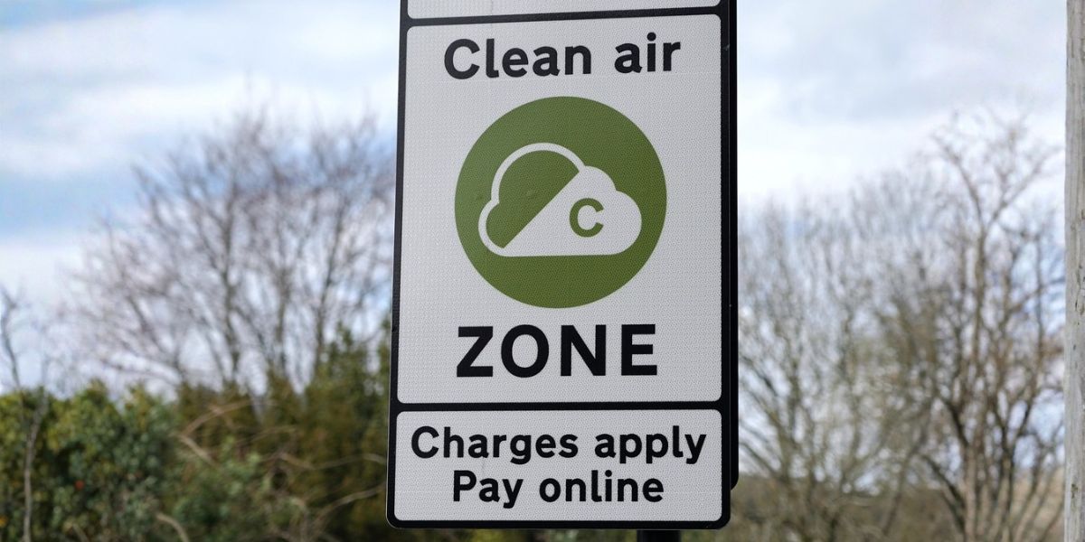 Fleet Clean Air Zone compliance: A white and black roadside sign reading "Clean Air Zone – Charges apply, Pay online" with the official green CAZ cloud logo, set against a backdrop of bare winter trees and a cloudy sky.