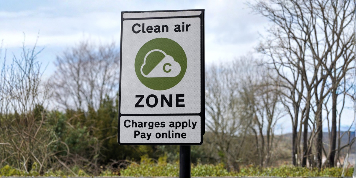 Fleet Clean Air Zone compliance: A white and black roadside sign reading "Clean Air Zone – Charges apply, Pay online" with the official green CAZ cloud logo, set against a backdrop of bare winter trees and a cloudy sky.