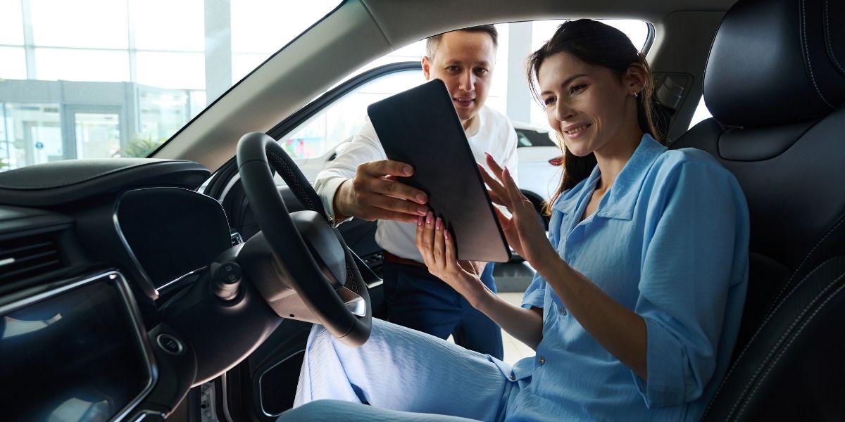 Fleet driver satisfaction A smiling woman seated in a car reviewing a tablet with a salesman leaning in through the open door beside her.