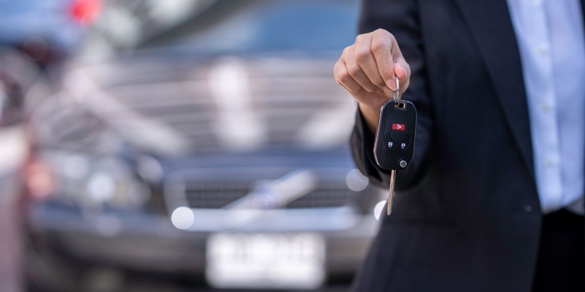 A person in a suit holding out a car key fob towards the camera, with a blurred dark car in the background.