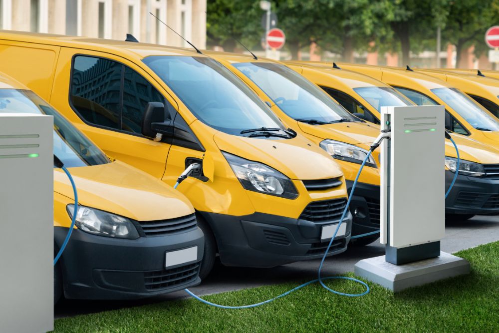 How to check if your fleet vehicles are CAZ compliant: A row of yellow electric vans parked and connected to EV charging points via blue cables in a fleet depot, with trees and buildings visible in the background.