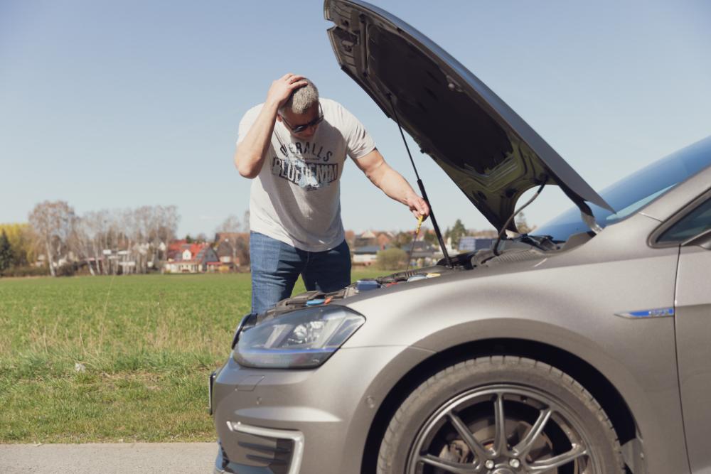 Pre-summer fleet checks for fleet managers UK: A frustrated man in a grey t-shirt and jeans holding his head with one hand whilst inspecting the open engine bay of a silver car at the roadside on a sunny day.