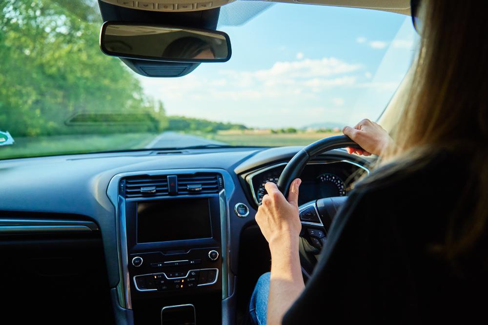 Preparing your fleet for summer high mileage: A view from the back seat of a right-hand drive car, showing a woman's hands on the steering wheel as she drives along a sunny rural road with green trees visible through the windscreen.