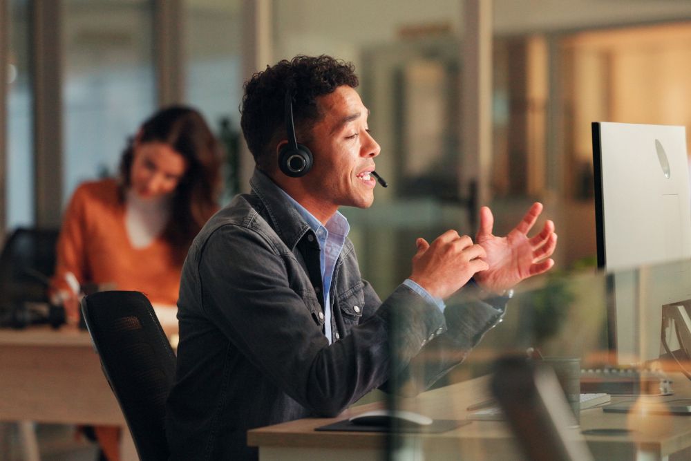 SG Fleet places to work employees SG Fleet places to work employees: A man wearing a headset speaks animatedly at his desk in a modern open-plan office, with a colleague working in the background.