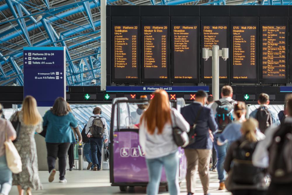The real cost of commuting for UK employees 2026: Commuters walking through a busy UK railway station concourse towards platform barriers, with a large departure board overhead displaying train times to destinations