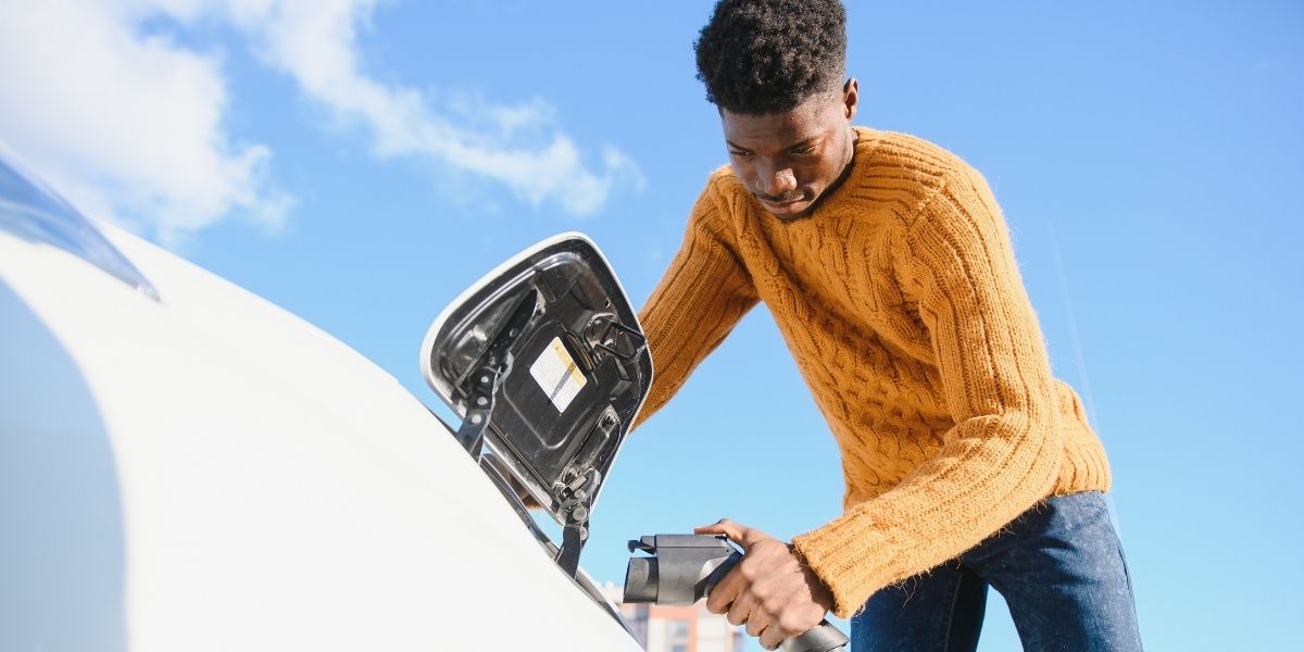 Vehicle fleet choices for staff A man in a mustard yellow jumper plugging a charging cable into the charge port of a white electric car beneath a blue sky.
