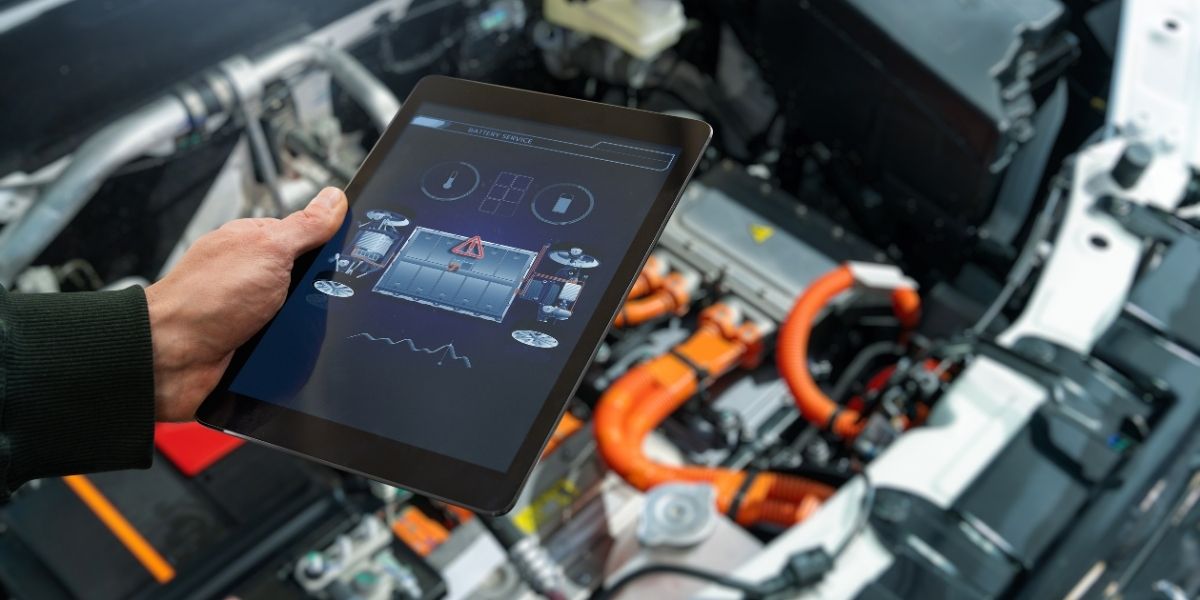 What reduces fleet operating costs beyond vehicles A hand holding a tablet displaying a battery service diagnostic interface, with a blurred electric vehicle engine bay showing orange high-voltage cables in the background.